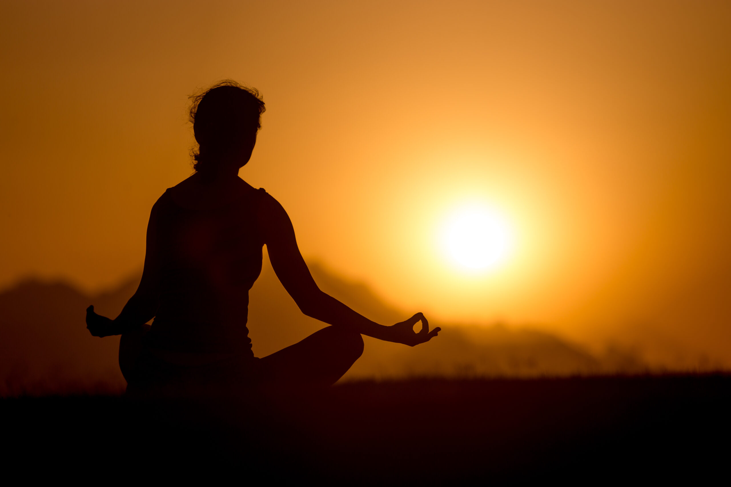 Silhouette of young woman sitting in picturesque serene place with crossed legs in yogic posture, looking at sunset or sunrise in mountains